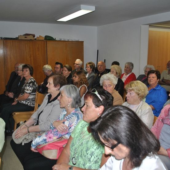 Ein Raum voller älterer Menschen, die auf Stühlen sitzen und einer Präsentation zuhören, mit einem hölzernen Schrank und Deckenbeleuchtung im Hintergrund.