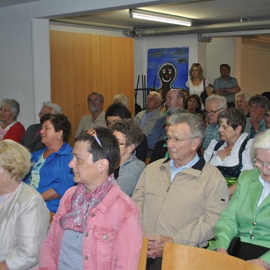 Eine Gruppe älterer Menschen sitzt in einem Besprechungsraum. Einige tragen Brillen, Halsketten und Ohrringe. Eine Frau in der Mitte trägt einen rosa Schal und eine rosa Jacke. Ein Mann vorne trägt eine beige Jacke. Der Raum hat eine Holztür, eine weiße Wand und ein blaues Gemälde an der Wand.