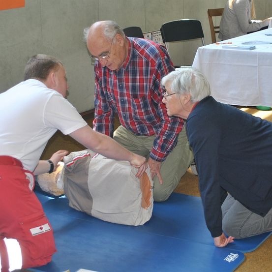 Drei Personen demonstrieren CPR an einem Mannequin auf einer blauen Matte. Ein Mann in einem roten karierten Hemd kniet und führt einen anderen Mann in einem weißen Hemd. Eine ältere Frau in einem marineblauen Pullover kniet in der Nähe.