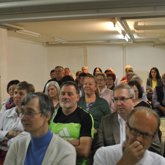 Eine Gruppe von Menschen sitzt in einem Raum und schaut nach vorne. Eine Frau sitzt in der vordersten Reihe, trägt eine weiße Strickjacke, und neben ihr sitzt ein Mann mit Brille und einem blauen Hemd. Ein Mann sitzt in der vordersten Reihe, trägt eine grüne Weste mit dem Adidas-Logo.
