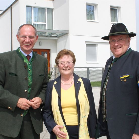 Drei Menschen stehen vor einem weißen Gebäude. Zwei Männer tragen grüne und schwarze Anzüge mit Krawatten, und eine Frau trägt ein gelbes Shirt und eine gelbe Jacke. Sie alle lächeln.