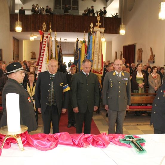 Vier Männer in Uniformen, darunter Militärkleidung, stehen um einen Tisch mit einem Kreuz, Kerzen und roten Bändern herum. Hinter ihnen steht eine Menschenmenge, von denen einige Hüte und Brillen tragen. Im Hintergrund befinden sich Fahnen und eine Galerie mit Menschen, möglicherweise Musikern.