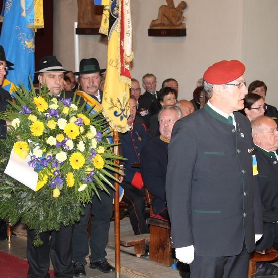 Eine Gruppe von Männern in Uniform und Mützen steht um einen Blumenstrauß in einem Raum. Einige sitzen auf Holzbänken, während andere stehen. Ein Mann trägt eine rote Mütze und eine Brille.