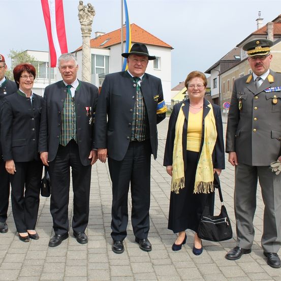 Eine Gruppe von Menschen steht auf einem gepflasterten Boden vor einem Gebäude mit Flagge und Statue. Sie sind in formeller Kleidung gekleidet, einschließlich Hüten und Schuhen.