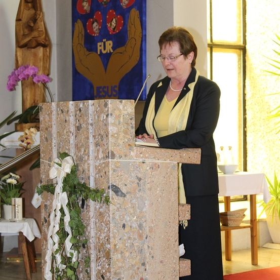 Eine Frau steht an einem Rednerpult mit einem Mikrofon und liest aus einem Buch. Hinter ihr ist ein blauer Banner mit dem Text 'FUR JESUS' zu sehen, und eine Statue einer Frau, die ein Kind hält, ist sichtbar. Der Raum ist hell beleuchtet.