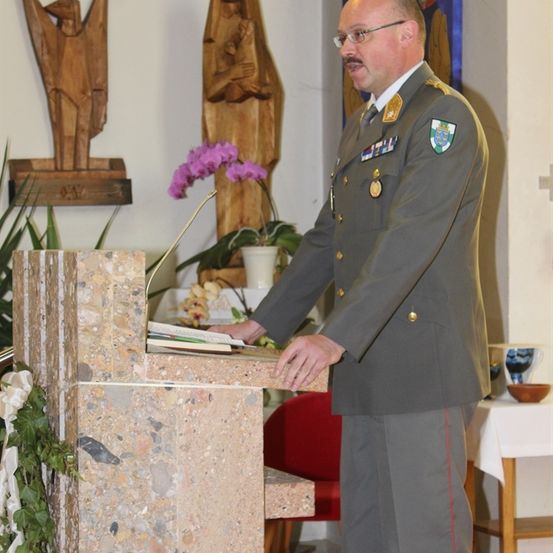 Ein Militäroffizier steht mit Büchern an einem Podest in einer Kirche mit einer Statue und Pflanzen im Hintergrund.