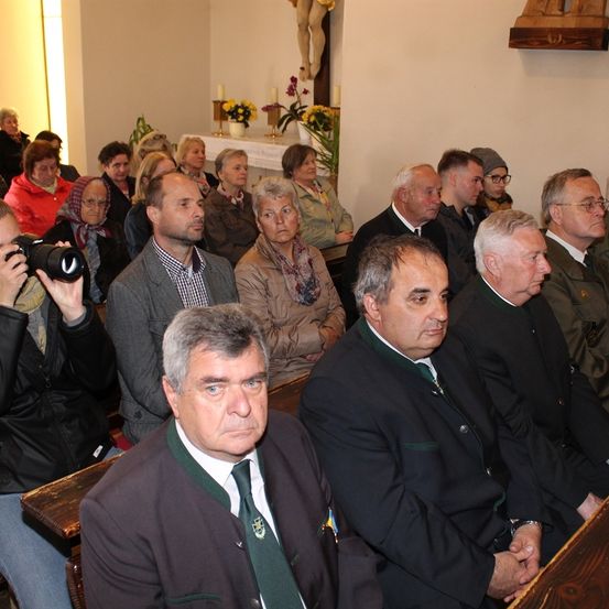Eine Gruppe von Menschen sitzt auf Kirchenbänken in einer Kirche, mit mehreren älteren Männern und Frauen in formeller Kleidung. Im Vordergrund hält ein Mann mit einer grünen Krawatte eine Kamera.