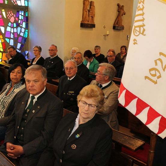 In einer Kirche sitzen mehrere Menschen, darunter Männer und Frauen, einige mit Brille und Anzug. Der Mann vorne hat eine grüne Krawatte, und die Frau vorne eine Brille. Hinter ihnen ist eine Wand mit einem Buntglasfenster und einer Flagge. An der Wand befinden sich auch Statuen einer Frau und eines Mannes.