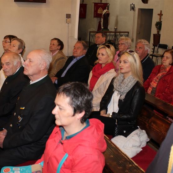 Eine Gruppe älterer Menschen sitzt in einer Kirche. Sie schauen auf etwas vor sich. Einige von ihnen tragen Brillen. Die Kirche hat eine Statue, ein Kreuz und einen Tisch mit einer Pflanze oben.