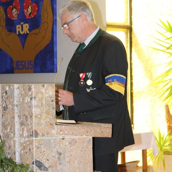 Ein Mann mit Medaillen an seiner Jacke liest von einem Buch an einem Podest in einer Kirche.