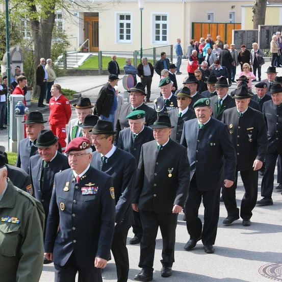 Eine Gruppe von Männern in Uniform, möglicherweise eine Militär- oder Polizeiparade, geht in einer Reihe vor einem Gebäude, hinter dem mehrere Menschen stehen.