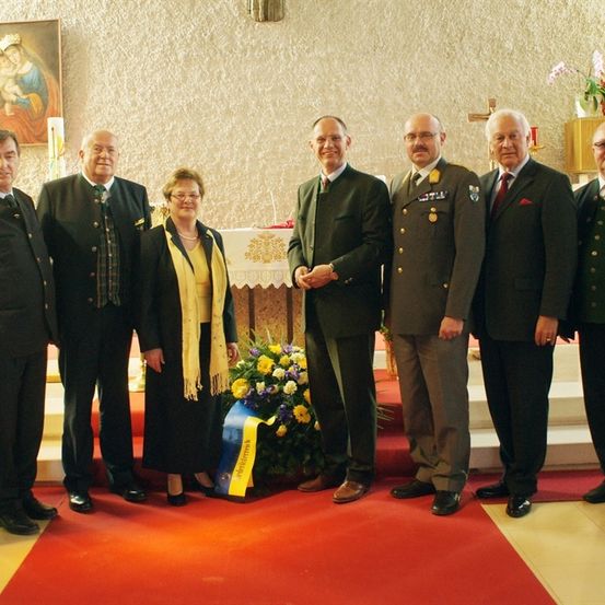 Eine Gruppe von Menschen in formeller Kleidung, einschließlich Militäruniformen, steht auf einem roten Teppich in einer Kirche. In der Mitte befindet sich eine Blumenarrangement, und ein Gemälde der Jungfrau Maria schmückt die Wand hinter ihnen.
