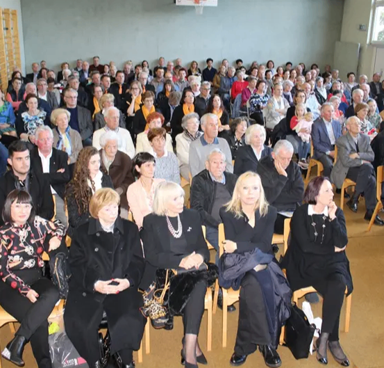 Eine Menschenmenge, bestehend aus Männern und Frauen, sitzt auf Stühlen in einem Raum. Einige stehen und unterhalten sich.