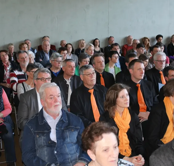 Eine große Zuhörerschaft, meist in formeller Kleidung, sitzt in einem Saal. Sie verfolgen eine Präsentation oder Veranstaltung. Das Publikum besteht aus Männern und Frauen, und einige tragen Brillen und Schals.