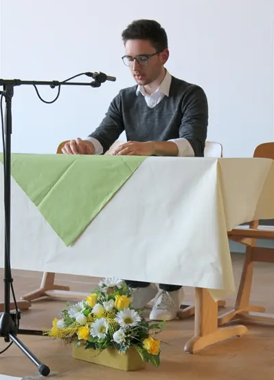 Ein Mann hält eine Rede auf der Bühne mit einem Mikrofon, trägt eine Brille und ein graues Langarmshirt. Vor ihm steht ein Tisch mit weißer Tischdecke und grüner Serviette sowie einer Blumenarrangement in einer Holzbox.