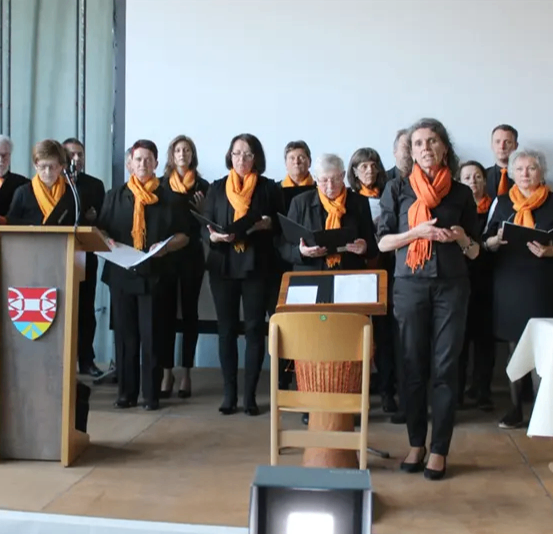 Ein Chor aus Frauen und Männern in schwarzen und orangefarbenen Schals steht auf der Bühne und liest von einem Notenständer, vor ihnen steht ein Rednerpult.