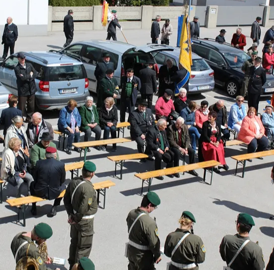 Men und Frauen sitzen auf Bänken in einem Kreis mit einer Blaskapelle in der Mitte. Hinter ihnen stehen viele Menschen. Mehrere Autos sind um sie herum geparkt.