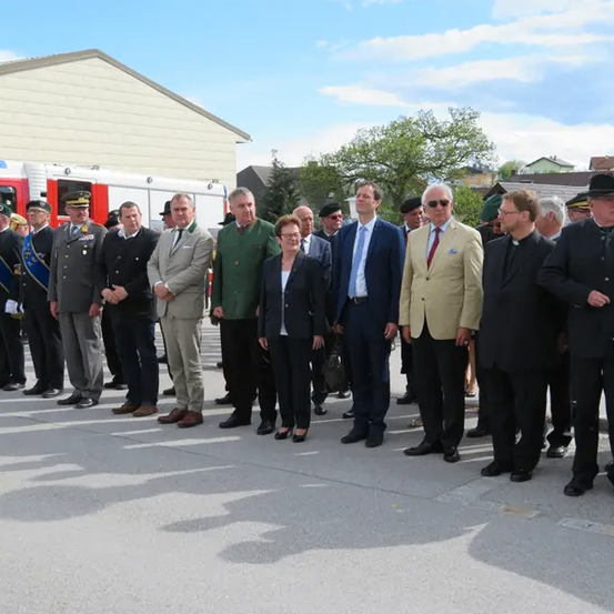 Eine Gruppe von Menschen in formeller Kleidung, darunter Anzüge und Uniformen, steht in einer Reihe auf der Straße, möglicherweise für eine Veranstaltung.