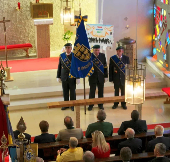 Drei Männer in Uniform stehen auf einer Bühne in einer Kirche. Ein Banner und eine Lampe stehen auf der Bühne. Dahinter befindet sich eine Wand mit einem Buntglasfenster. Das Publikum sitzt auf den Kirchenbänken.