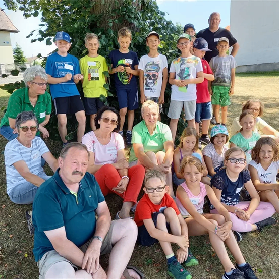 Eine Gruppe von Kindern und Erwachsenen, darunter zwei Frauen und ein Mann, sitzen und stehen auf einem Rasen und lächeln für ein Foto.