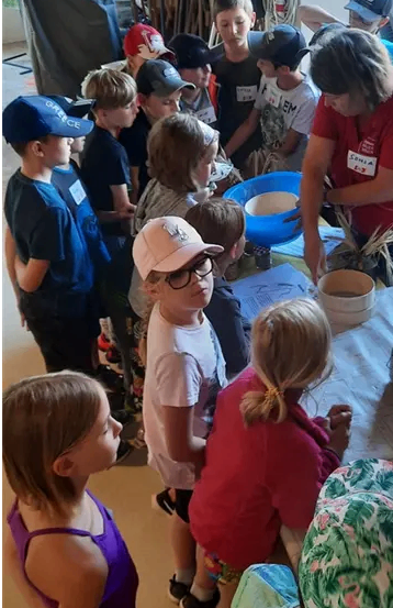 Eine Gruppe von Kindern und zwei Erwachsenen versammelt sich um einen Tisch. Die Erwachsenen bereiten Essen in Schüsseln zu. Einige Kinder tragen Hüte und Brillen.
