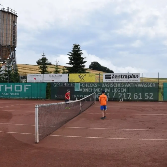 Zwei Personen spielen Tennis auf einem Platz mit einem Netz und Bannern dahinter, darunter eines für Zentraplan.