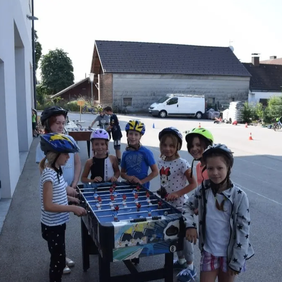 Eine Gruppe von Kindern mit Helmen steht vor einem Tischkicker in einer Einfahrt.