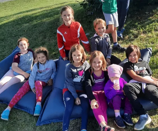 Eine Gruppe von Kindern sitzt auf blauen Bohnensäcken auf einer Wiese. Ein Kind hält einen Becher.