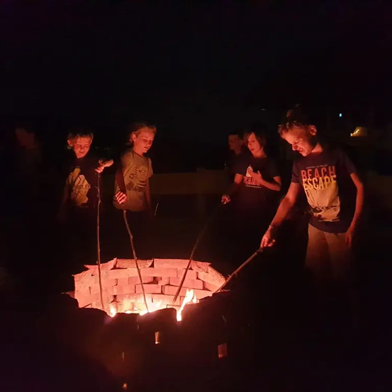 Fünf Kinder stehen um ein Lagerfeuer mit Holzstäben herum. Drei von ihnen halten Stöcke, während die anderen zwei Stöcke mit Marshmallows halten.
