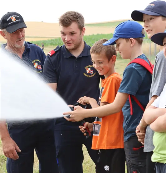 Zwei Feuerwehrleute in Uniform stehen mit drei Jungen. Ein Junge sprüht Wasser aus einem Schlauch.