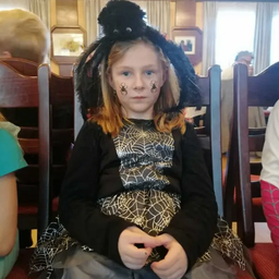 Ein kleines Mädchen sitzt auf einem Stuhl, gekleidet in ein Halloween-Kostüm mit Spinnweb-Designs und hat Spinnweben auf ihrem Gesicht gemalt.