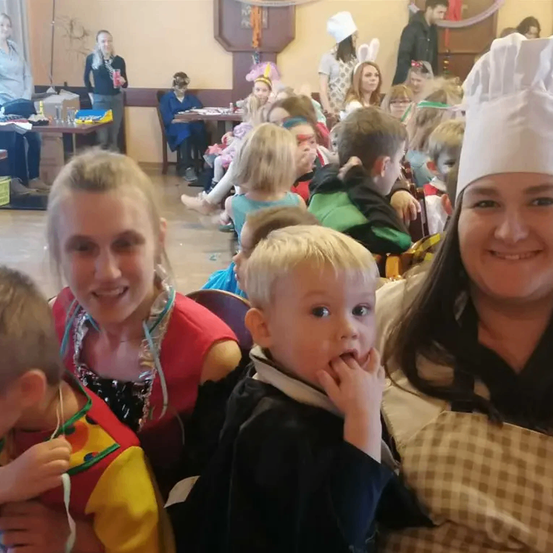 Eine Gruppe von Menschen, darunter Kinder in Kostümen, sitzen in einem Raum. Eine Frau in einer Kochmütze lächelt und posiert für ein Foto mit einem Jungen und einem Mädchen.
