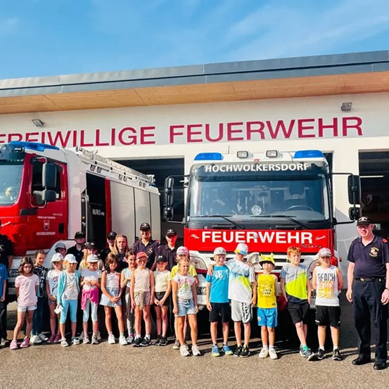 Gruppenfoto von Kindern mit Feuerwehrleuten vor einer Feuerwache mit roten Feuerwehrwagen