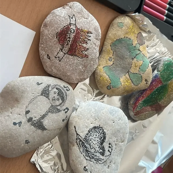 Fünf handbemalte Steine auf einem Schreibtisch, jeder mit einzigartigen Designs. Ein Stein hat eine Krone, ein anderer eine grüne Landschaft und andere haben Musiknoten, einen Engel und eine Krone.