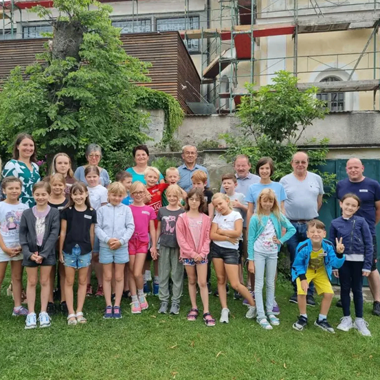 Eine Gruppe von Kindern und Erwachsenen steht auf einer Rasenfläche vor einem im Bau befindlichen Gebäude.