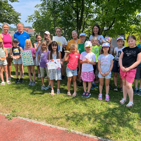 Eine Gruppe von Kindern und Erwachsenen steht auf einem Rasen, einige halten Gemälde in den Händen und tragen Hüte, Brillen und Turnschuhe.