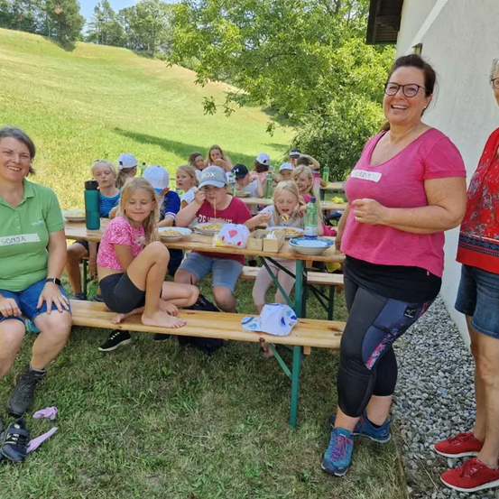 Eine Gruppe von Menschen, darunter Frauen und Kinder, sitzt an einem Picknicktisch. Einige essen, andere schauen zu. Sie befinden sich auf einer Wiese, umgeben von Bäumen.