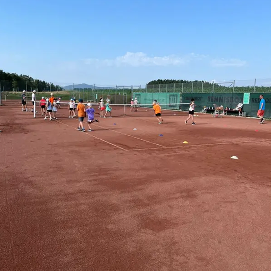 Eine Gruppe von Kindern spielt Tennis auf einem Sandplatz. Sie tragen Turnschuhe und Mützen, manche haben Tennisschläger dabei. Der Platz hat ein Netz in der Mitte und ist von einem Zaun umgeben. In der Ferne gibt es Bäume und Berge.