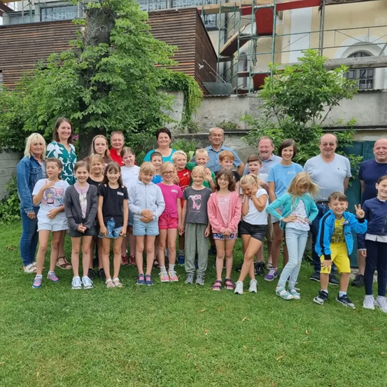 Eine Gruppe von Menschen steht in zwei Reihen in einem Garten, lächelt und posiert für ein Foto. Einige tragen eine Brille, Turnschuhe und manche haben Armbänder an den Handgelenken.