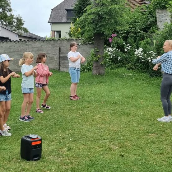 Eine Frau steht in einem Garten mit fünf Kindern. Sie hören alle Musik von einem Lautsprecher.