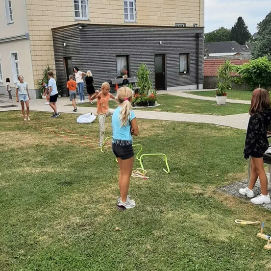 Eine Gruppe von Kindern spielt mit einem gelben Hula-Hoop auf einer Rasenfläche vor einem Gebäude.