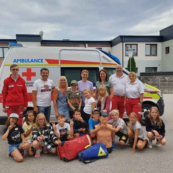 Eine Gruppe von Menschen posiert vor einem Krankenwagen. Einige von ihnen tragen Mützen, Brillen und Turnschuhe. Ein Mann hält einen Dummy und steht vor dem Krankenwagen.