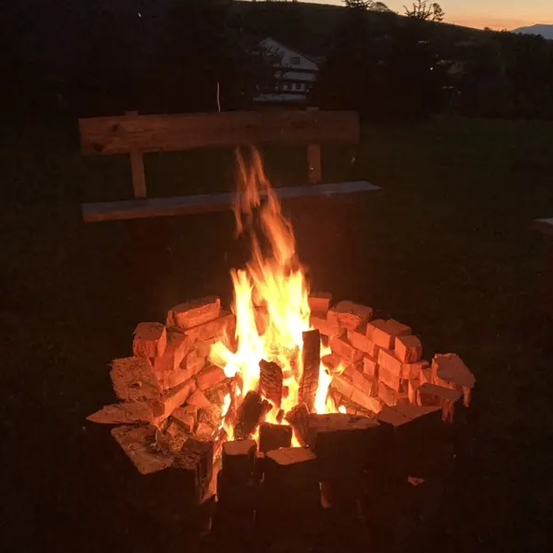 Ein Lagerfeuer brennt am Abend hell, umgeben von Holzscheiten und neben einer Holzbank auf einem Grashügel.