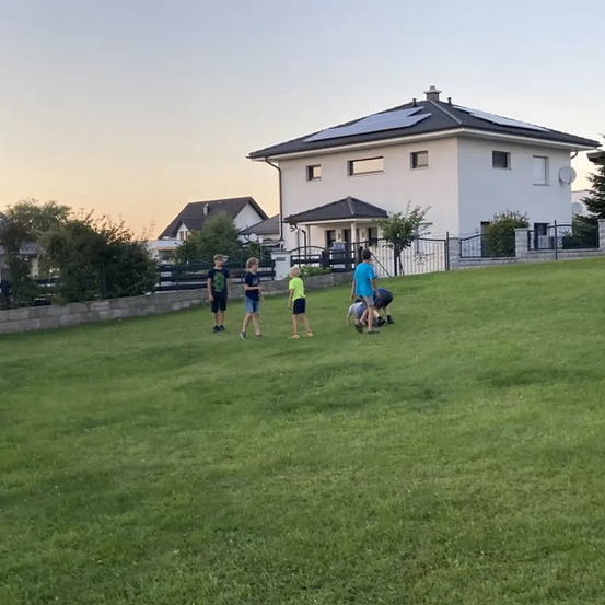 Fünf Kinder spielen auf einem Rasen vor einem modernen Haus mit Solarpanelen auf dem Dach.