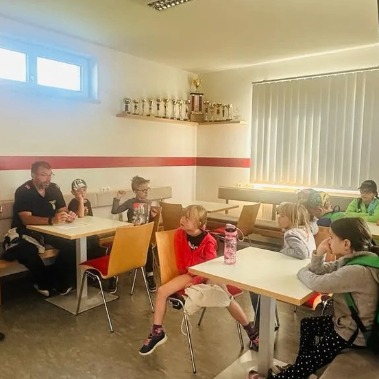 Eine Gruppe von Kindern sitzt in einem Raum, wahrscheinlich einem Klassenzimmer, an Tischen. Der Mann, der vor ihnen steht, ist wahrscheinlich der Lehrer. Der Raum hat einen Tisch und Stühle, ein Regal mit Trophäen und eine Tafel mit einem Vorhang.