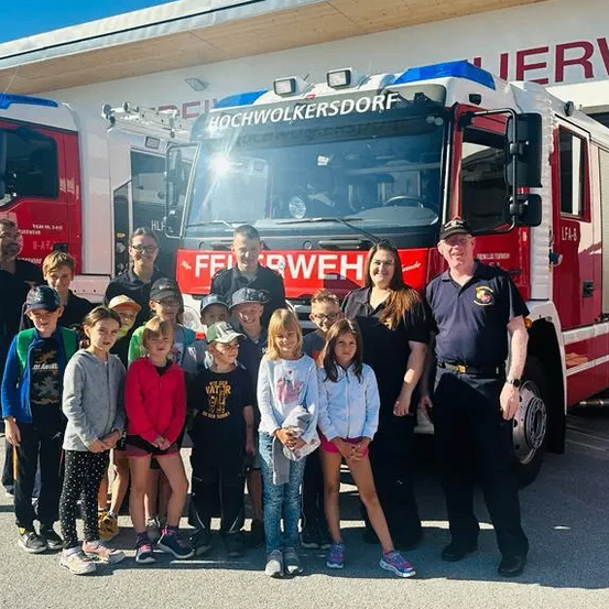Eine Gruppe von Kindern und Erwachsenen steht vor zwei roten Feuerwehrwagen, wobei zwei Männer in Uniformen vor einem der Wagen stehen.