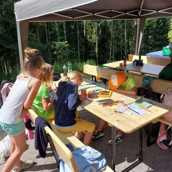 Vier Kinder sitzen unter einem Zelt an einem Tisch und malen. Ein Kind trägt einen Hut und scheint zu schlafen.
