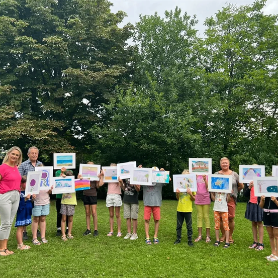 Eine Gruppe von Kindern und zwei Erwachsenen steht auf einer Rasenfläche und hält Zeichnungen in den Händen. Die Zeichnungen haben bunte Illustrationen und werden vor den Kindern gezeigt. Die Erwachsenen stehen an den Seiten der Kinder. Hinter ihnen stehen Bäume mit üppigen grünen Blättern.