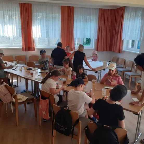 Ein Klassenzimmer mit Kindern, die an langen Tischen sitzen, einer Frau, die vorne steht, und einer anderen Frau, die an einem Tisch sitzt.
