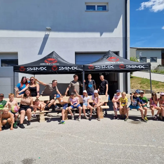 Eine Gruppe von Menschen in Freizeitkleidung sitzt auf Holzbänken unter einem Zelt, mit zwei Zelten dahinter, auf denen das Wort '24MX' steht.